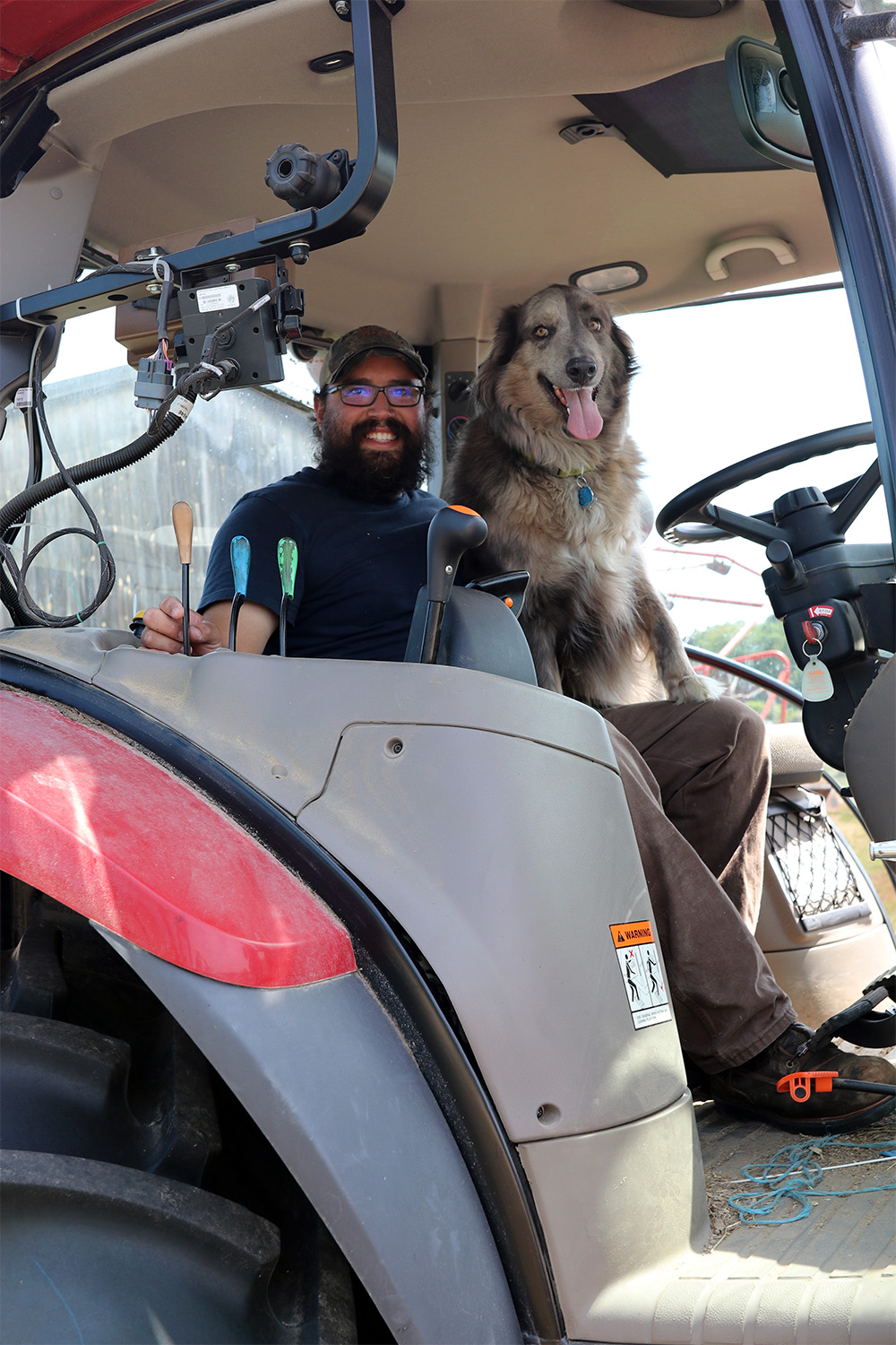 A photo of a white male with a blue shirt, brown pants and a hat sitting in a tractor cab next to a grey, white and brown dog.