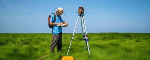 A student with surveying equipment, wearing a hat, stands in a sea of grass.