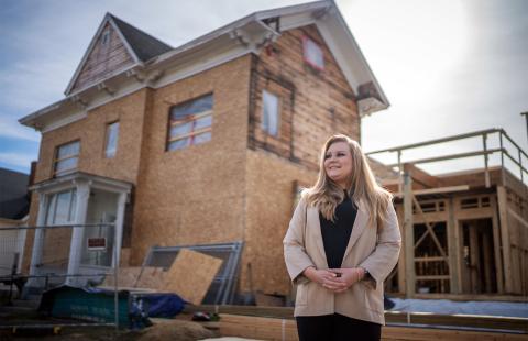 A photo showing COLSA alum Cassie Mullen '16, a white woman with long blond hair, standing in front of a two-story building that is under construction. Cassie looks off in the distance.