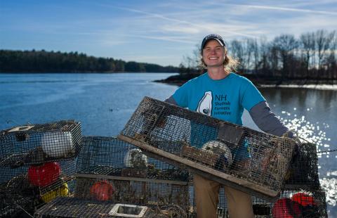 UNH COLSA alum Kelsey Meyer stands on a boat holding up a shellfish trap and wearing a t-shirt with the words NH Shellfish Farmers Initiative on it. Kelsey is white with hair tucked beneath a ballcap and the t-shirt is teal.