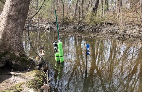 A water quality sensor attached to floats and a pole stuck into a muddy brown river surrounded by forests.
