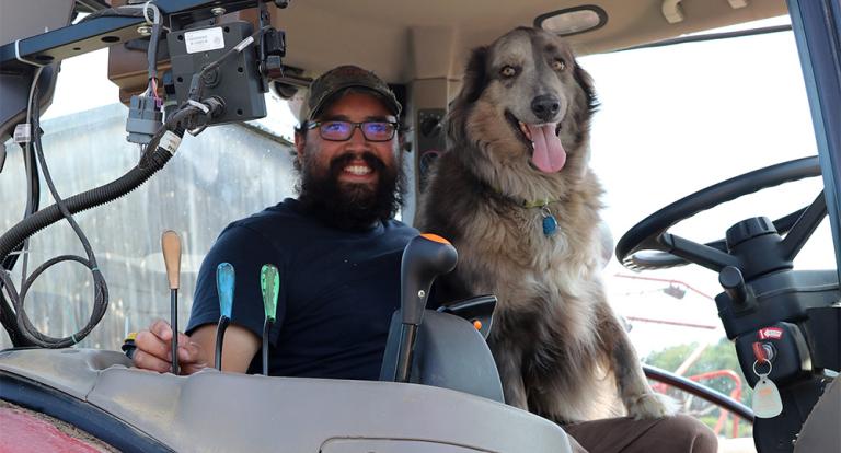 A photo of a white male with a blue shirt, brown pants and a hat sitting in a tractor cab next to a grey, white and brown dog.