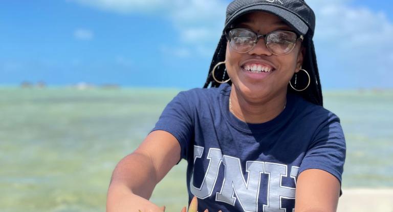A photo of a black woman with glasses wearing a blue UNH t-shirt, standing up in a boat in open water and holding out a clump of seaweed to the camera.