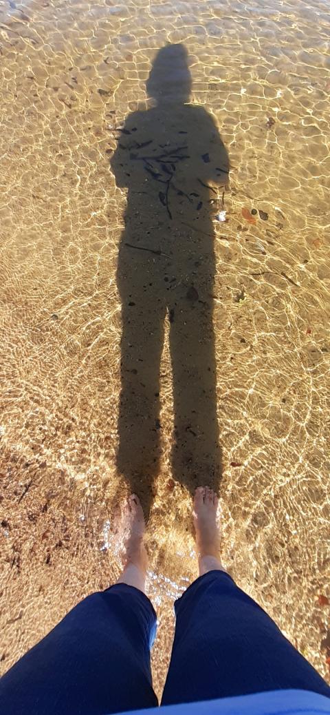 A person's legs and feet (wearing blue pants) visible in the foreground, their shadow outstretched before them in crystal-clear waters.
