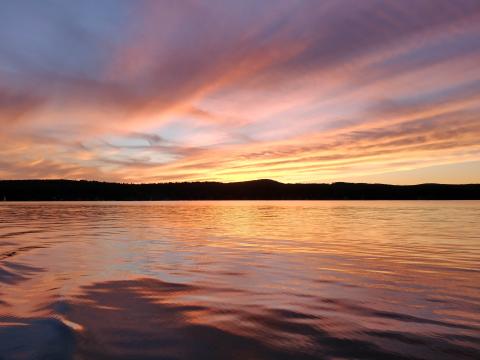 A sunset photo, the shoreline in the horizontal center of the photo, the water below and sky above. Sky is bluish, purplish, orange, reflecting in the water, turning it yellowish blueish. 