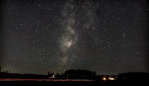 A nighttime photo showing tiny white dots–stars–speckling the sky. Near the bottom is a dark outline of trees and a few small areas of houselights. A clouded area in the vertical center of the photo shows the milky way.