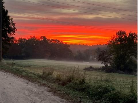 A photo taken of a sunrise that makes the top half of the frame–showing the sky–deep orange and yellow. In the bottom half of the frame, a field with mist upon it.