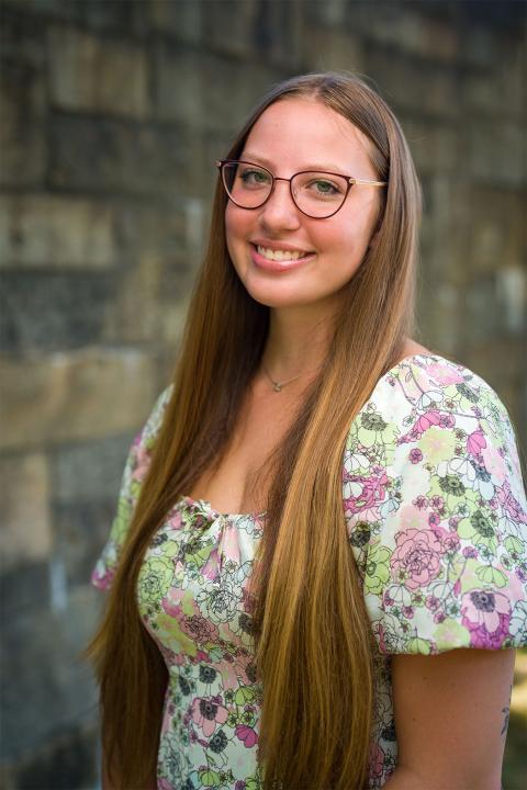 Master of Science in Nutrition student Amy Urban. Amy has white skin, long brown hair that reaches to her waist. She wears a purple, pink and green floral printed top. She wears glasses with brown rims.