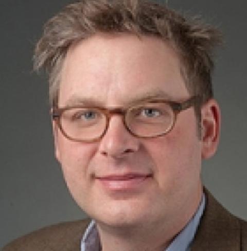 A photo of COLSA researcher David Plachetzki. David is a white man. In this photo, he wears brown-rimmed glasses, has brownish-blonde hair. He is half smiling and has a brown suit jacket on.
