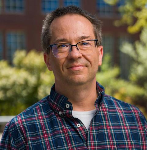 A photo of a white man, Matt MacManes, wearing glasses and a plaid shirt with a white undershirt. Matt is smiling while looking into the camera. Behind him is a blurred brick building with windows and vegetation.