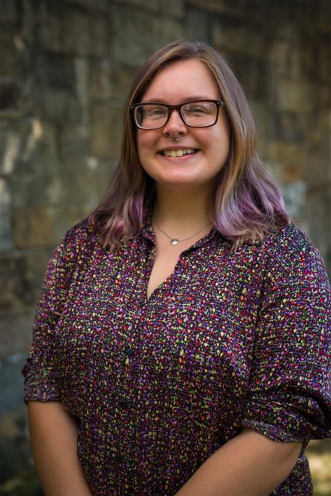 Master of Science in Nutrition student Rachel Goding. Rachel has brown hair with tinges of purple on the ends. She has white skin and wears brown-rimmed glasses. Her shirt is purple with dots of yellow, green and red.