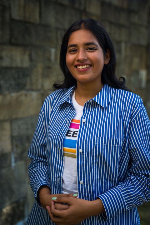 Master of Science in Agricultural Sciences student Radhika Rani. Radhika has dark-brown skin and jet-black hair. She wears an unbuttoned button-up shirt with blue and white stripes. Underneath the shirt she wears a colorful t-shirt.