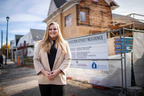 A photo showing COLSA alum Cassie Mullen '16, a white woman with long blond hair, standing in front of a sign that says "Street Residence" on a sign hung on a chainlink fence. Behind that is a building under construction.