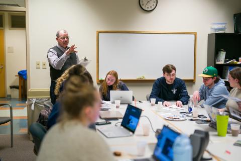 Instructor Drew Conroy, a while male with a balding head, stands in the background next to a whiteboard and speaks to a group of students seated at a table in front of him. The students work on laptops and notepads.