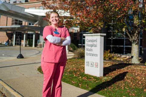 Medical laboratory science degree student Svetlana (Lana) Gerace ’22 stands near a sign stating "Concord Hospital Maine Entrance." She's dressed in pinkish-red scrubs.