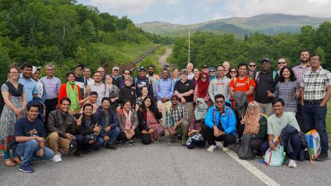 A photo of a large group of people standing in a road with mountains and forests in the background.