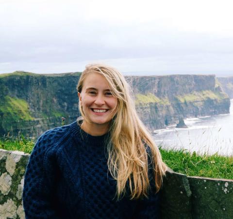 A photo of a white woman standing in front of a stone wall, large cliffs shown in the background