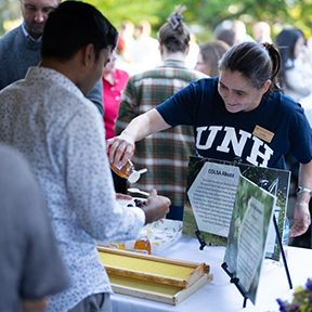 Person wearing UNH t-shirt and nametage pours a sample of honey for an event attendee