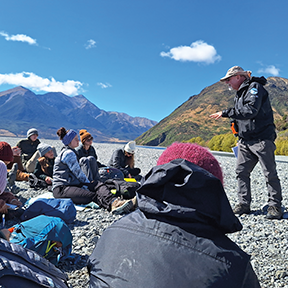 A group of students sitting on the ground outside in New Zealand listening to an instructor wearing a black jacket and hat