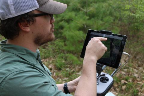 A white male wearing a baseball cap points to a screen he holds in front of him. He is in the forest.