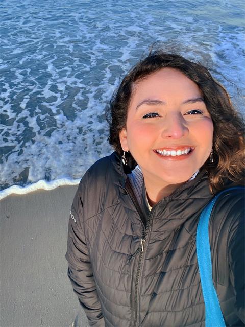 A photo of a woman smiling, holding the camera up in her left hand, at the beach and in front of the water.