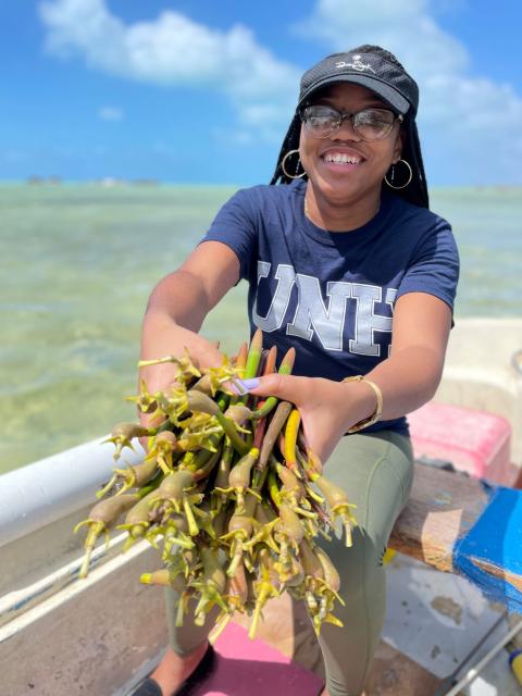 A photo of a black woman with glasses wearing a blue UNH t-shirt, standing up in a boat in open water and holding out a clump of seaweed to the camera.