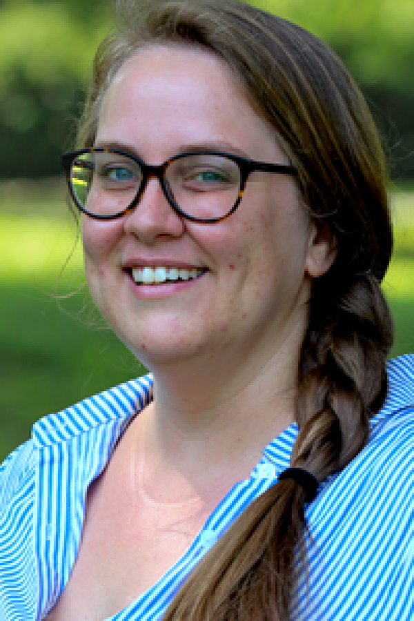 A photo of researcher Anna Wallingford. Anna is a white woman. She wears brown-rimmed glasses and has her hair in a ponytail. She wears a button shirt with blue and white stripes.
